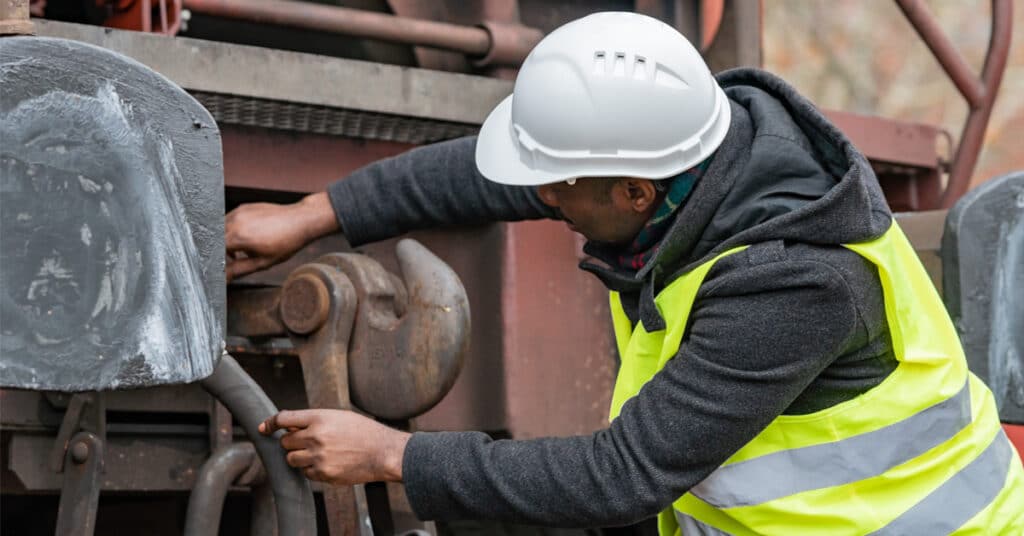 A railcar maintenance worker in a yellow vest and white safety hat inspects a railcar.