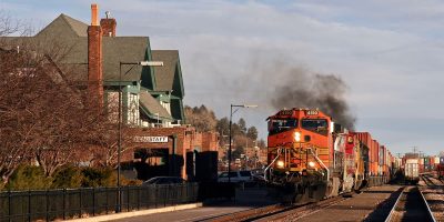 An orange and yellow rail freight train hauls rail cargo through a small town.