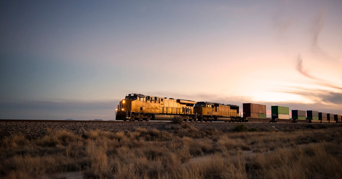 A yellow freight train hauls loads of colorful boxcars through a shrub desert against a setting sun.