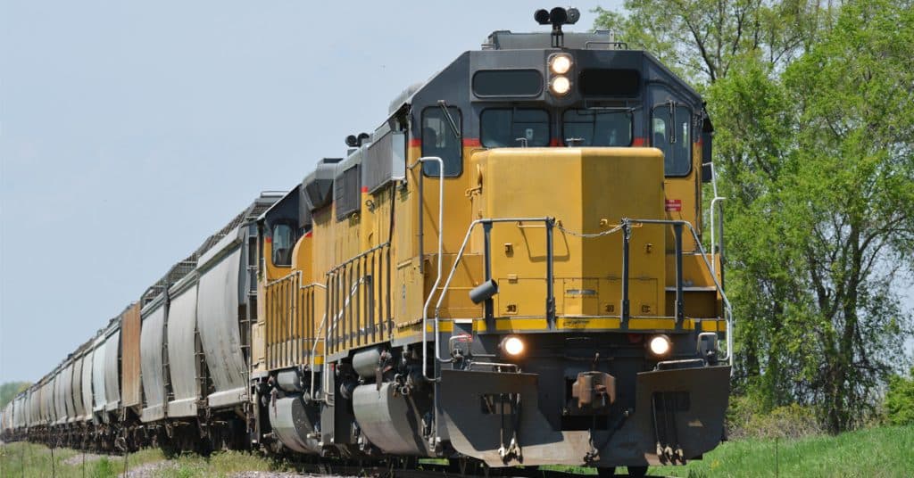 A yellow freight train hauls a line of rail cars through green grass and trees.