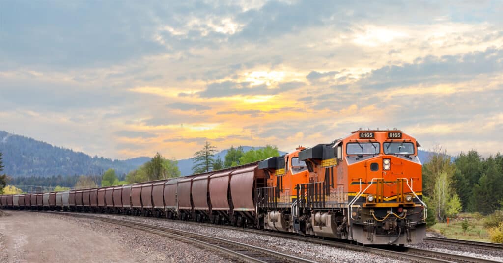 An orange train pulls a train of hopper rail cars through the desert with mountains in the backgrounds.
