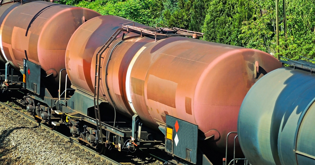 Three brown and grey oil tanker railcars along a track next to woods.