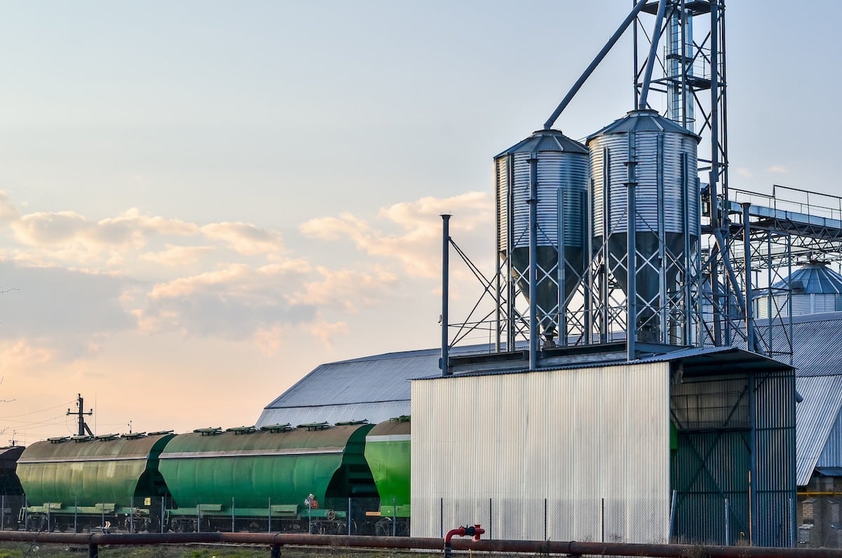 Green rail hopper cars lined up in front of a silver manufacturing factory.