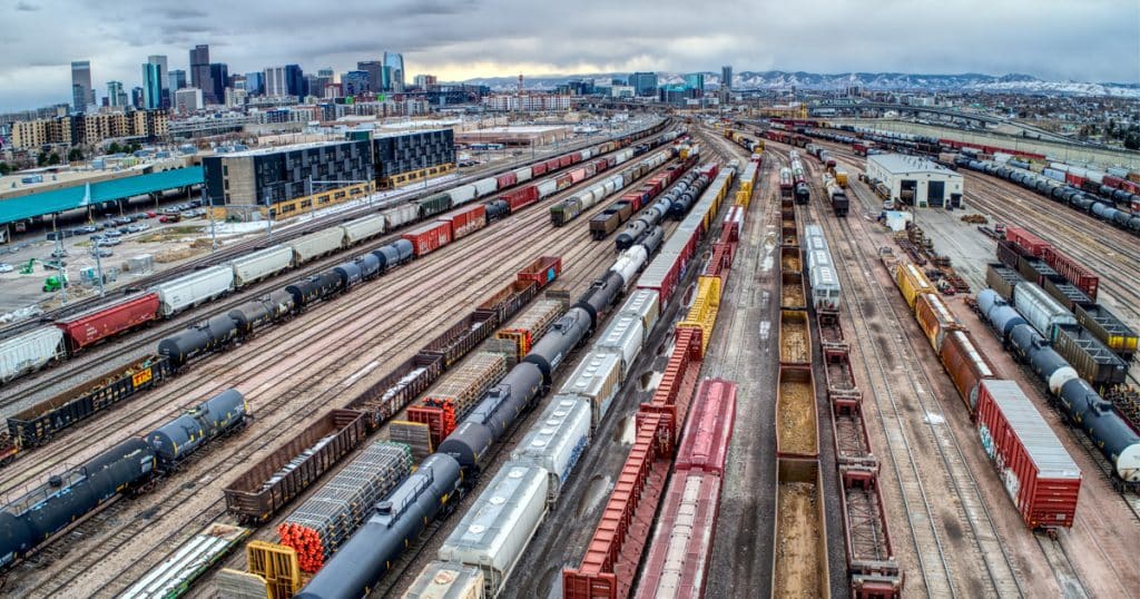 Lines of freight rail cars in a rail yard with the Denver, Colorado skyline in the distance.
