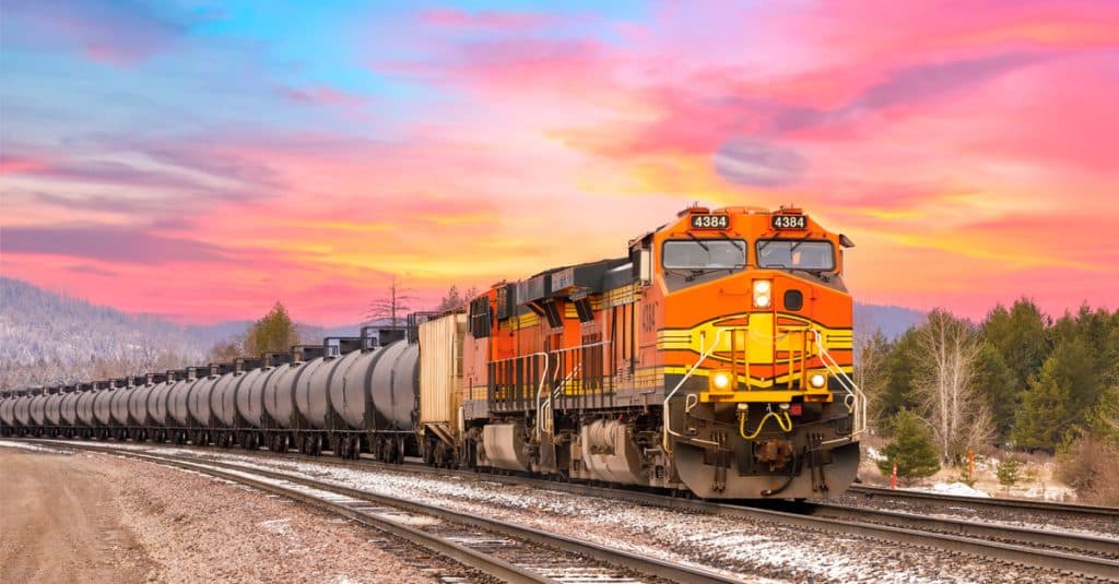 An orange rail locomotive hauls a line of black rail tank cars in front of a yellow and pink sunset.