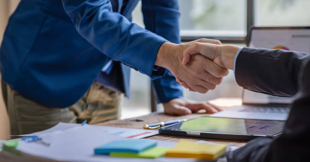Two business professionals shake hands over a table of laptops, tablets, and papers.