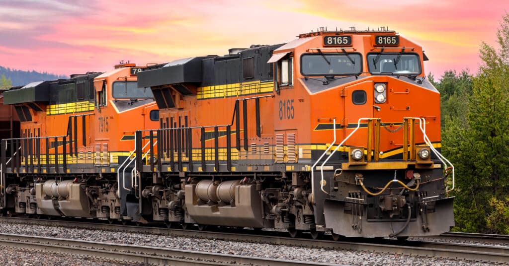 An orange rail freight engine hauls another orange engine in front of a sunset.