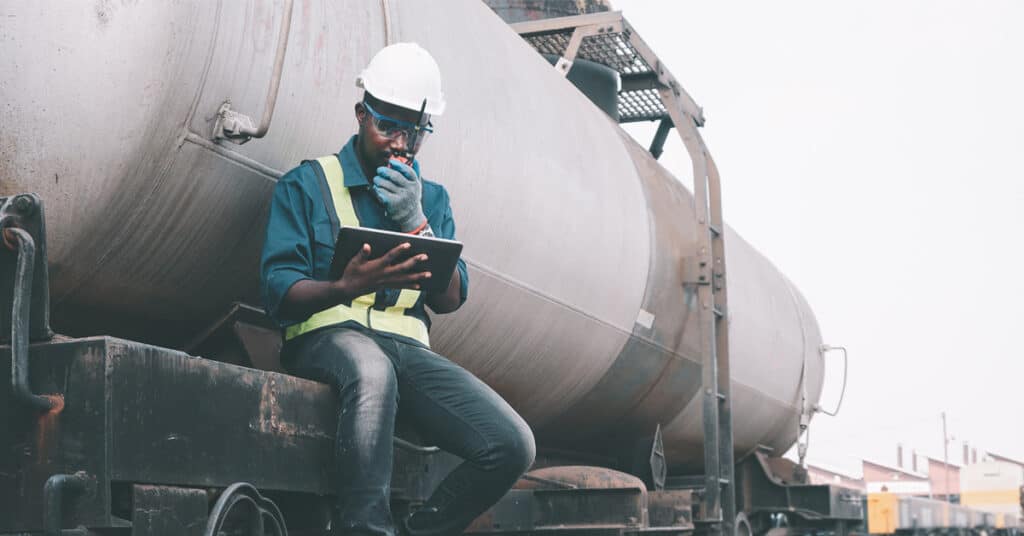 Train repair employee sits on a tankcar talking by radio communication while looking at a tablet.