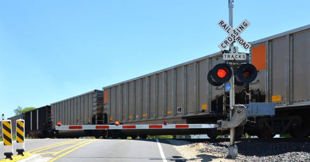 A line of grey rail hopper cars passes over a road with a red railroad crossing sign.