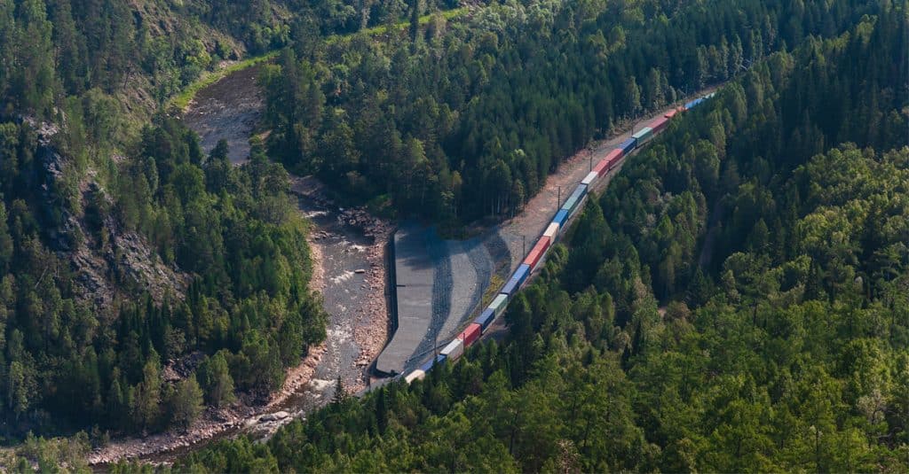 A top down view of a line of colored railcars amidst a green forest beside a stream.