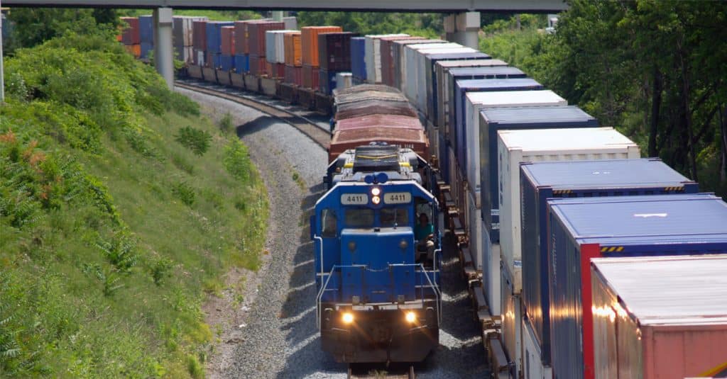 A blue rail engine pulls a few railcars alongside a long series of intermodal railcars.