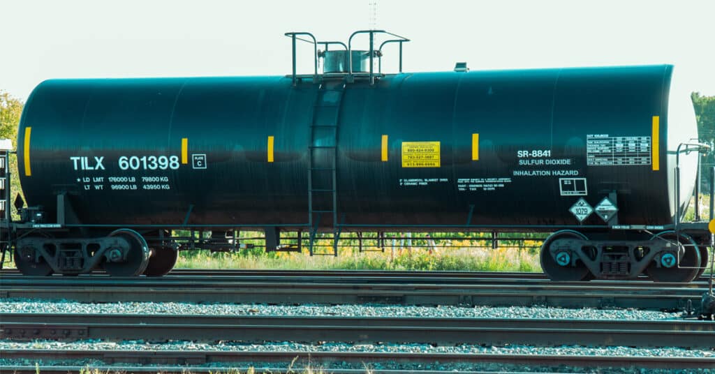 A black Trinity tank car with yellow stripes sits on a rail in a rail yard.