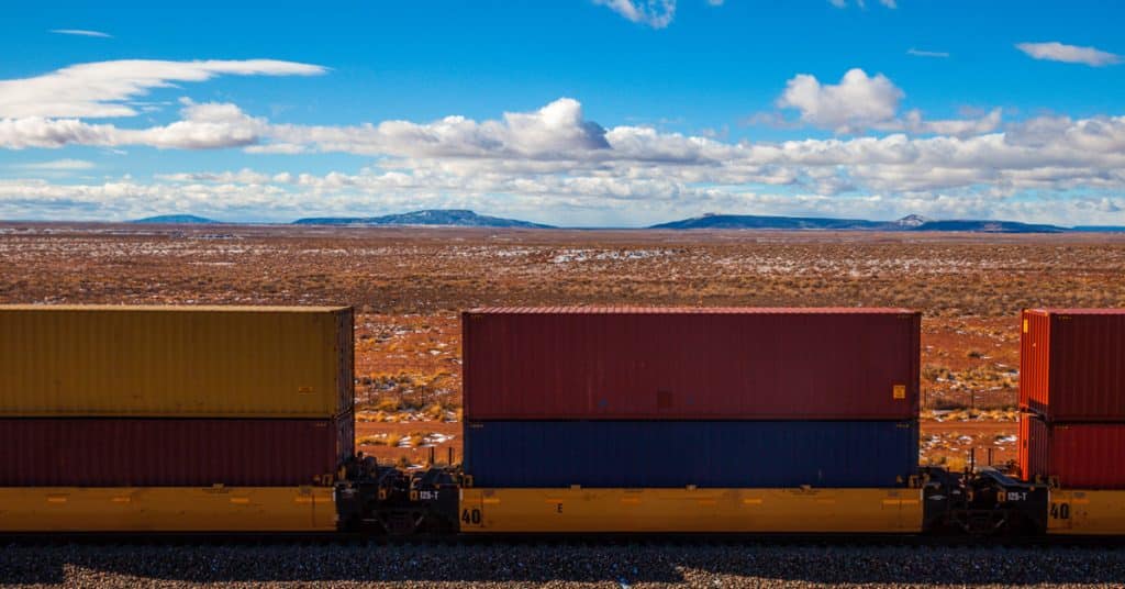 A row of double stacked rail cars travels across the desert with mountains on the horizon.
