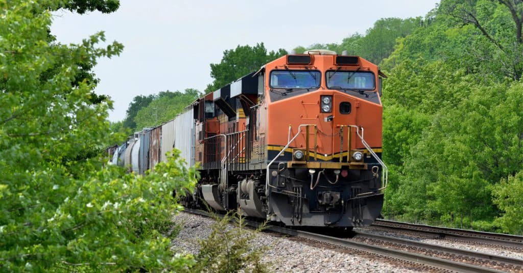 An orange engine hauls rail cars through green trees.