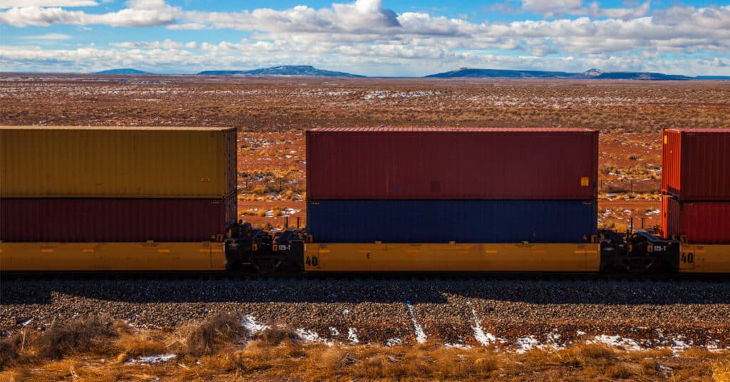 Colorful stacked intermodal railcars move on a rail track through a desert with mountains on the horizon.
