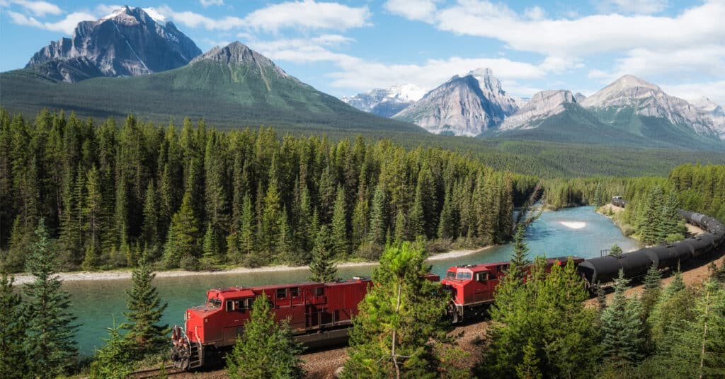 Red cargo train hauling black rail tank cars through a forest alongside a blue river with towering mountains in the background.