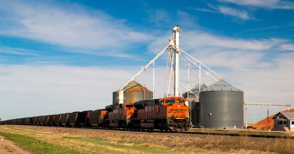 A freight train with an orange locomotive passes in front of a series of grain silos in a green plain.