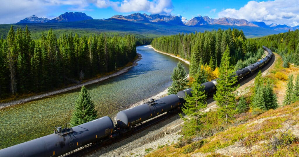 A train of rail freight tank cars moving through a forest along a river with mountains in the background.