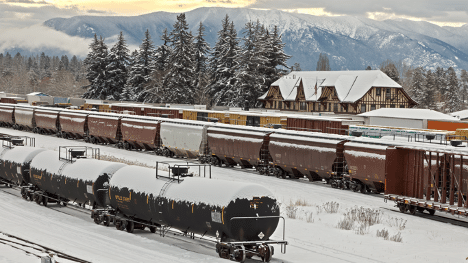 Montana rail yard and depot in winter with snow covered tank cars, rail cars, and mountains in the background.