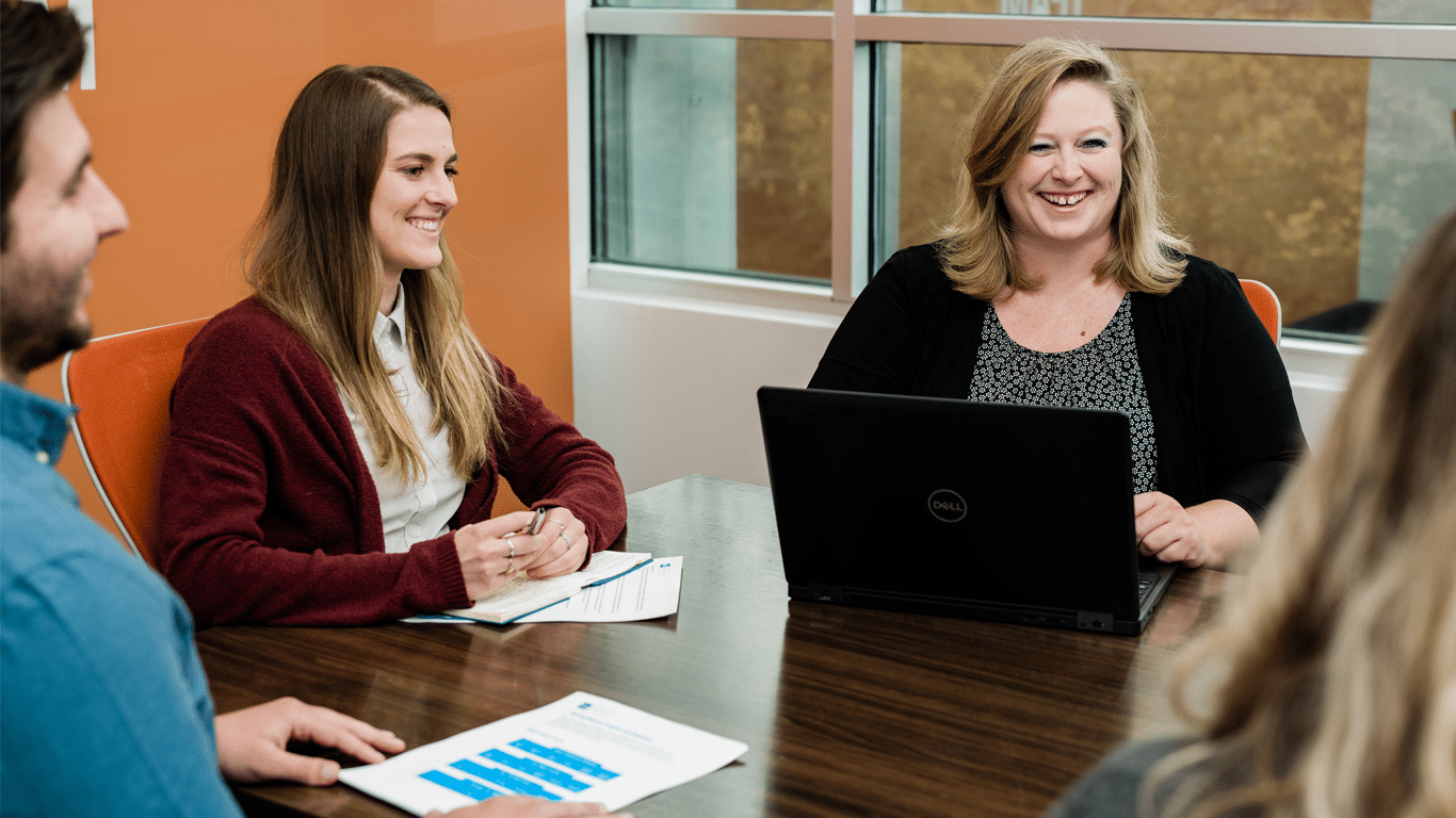 Three smiling RSI Logistics experts sit around a table with laptops and graphs in an orange conference room.